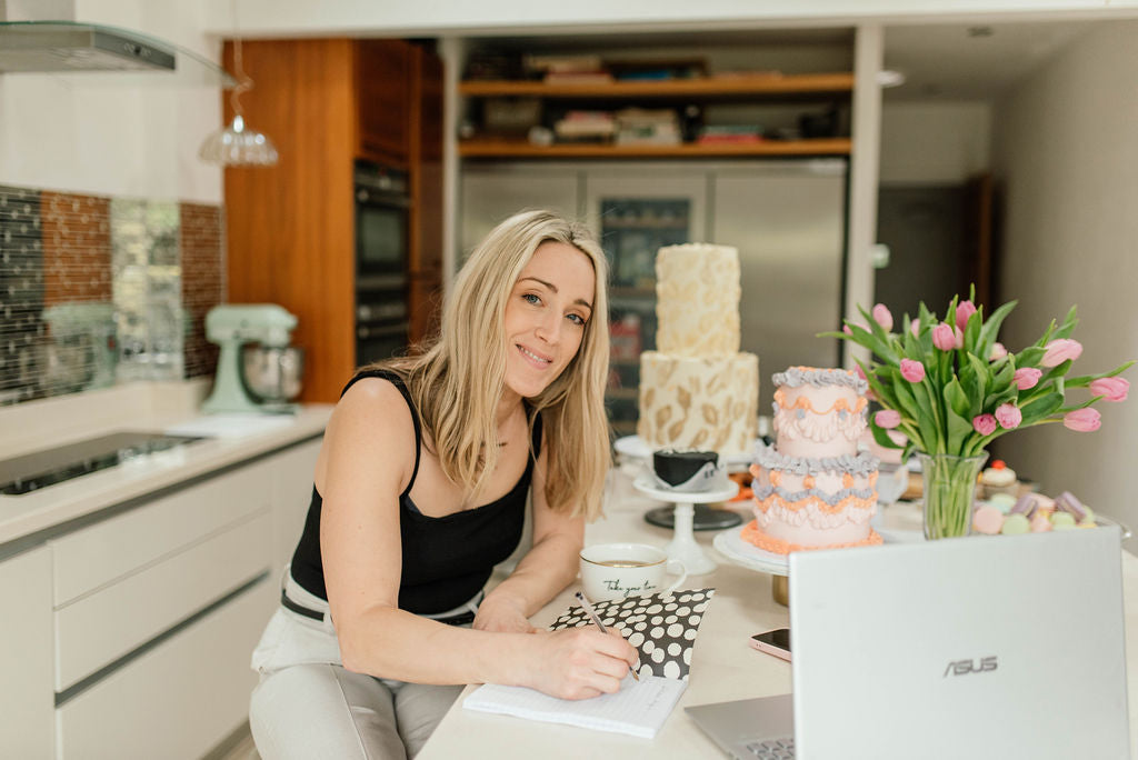 Baker Jo sitting and writing in notepad, beautiful celebration cakes and sweet treats sit on the counter behind her.